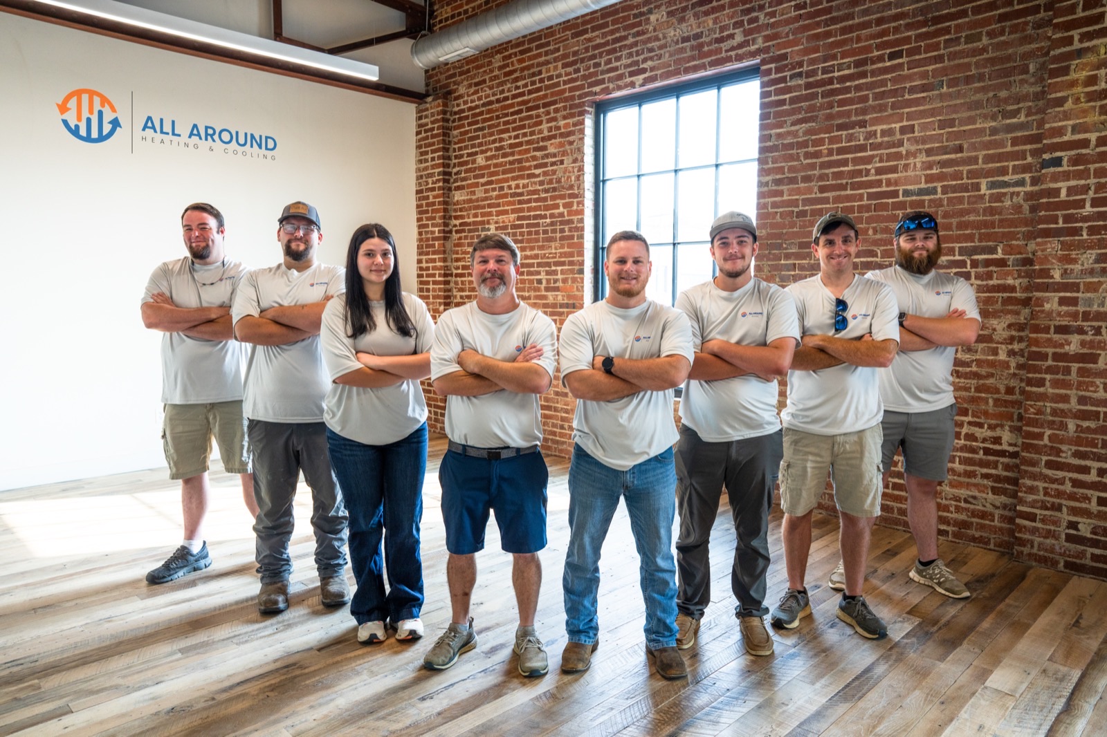 The eight-person All Around Heating & Cooling team in matching shirts, standing in front of the company logo wall at their Cullman, Alabama office